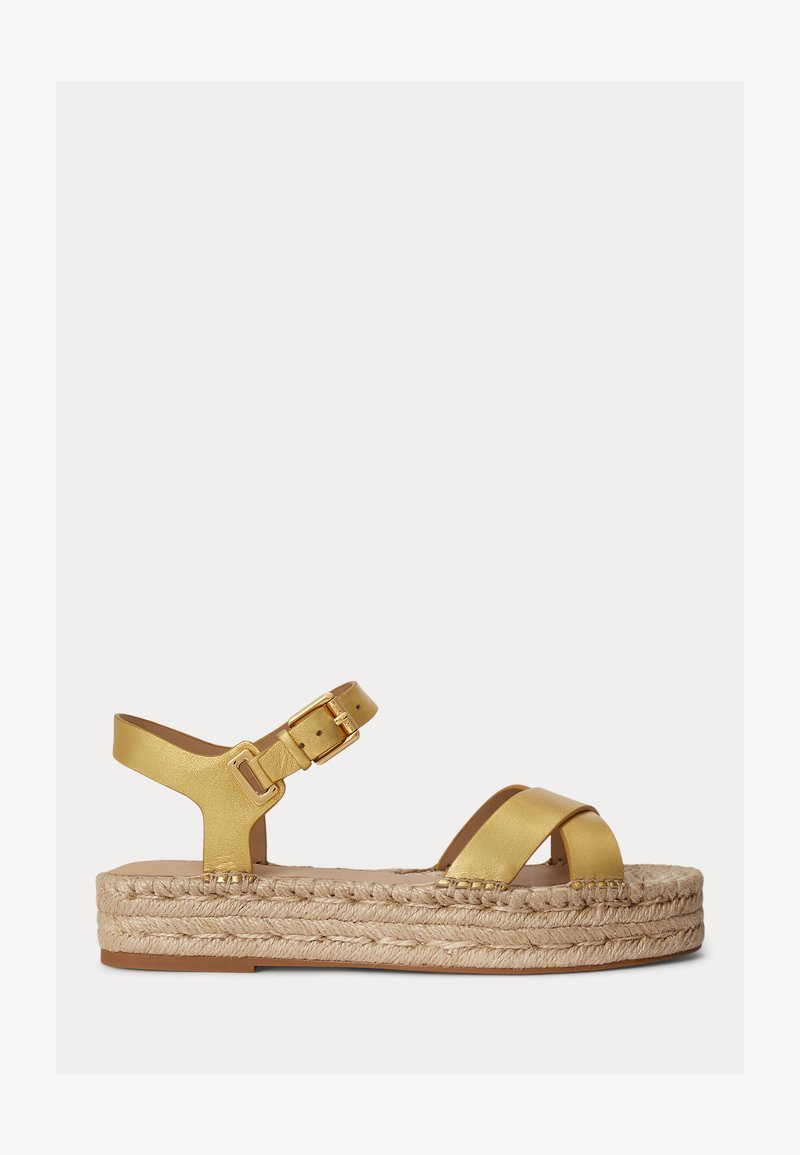 Guld læder kilehælssandal med ankelrem og spænde, flettet jutesål og krydsstropper over tæerne, sideudsigt på hvid baggrund.