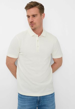 Uomo che indossa una polo bianca a maniche corte con tessitura e jeans blu, in piedi con le mani dietro la schiena, che guarda verso il basso di lato.