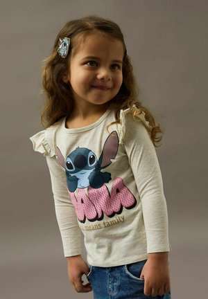 Beżowa koszula z długim rękawem z nadrukiem postaci z kreskówki „Stitch” w różowym kolorze, z falbanami na rękawach. Noszona z niebieskimi szortami dżinsowymi.