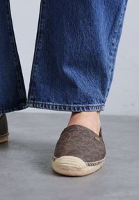 Soulier espadrille avec un dessus à motifs brun foncé, semelle en jute et bord tissé. Porté avec un pantalon en denim bleu large, affichant une coupe décontractée.