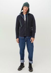 Dunkelblaue Reißverschluss-Fleecejacke mit Kapuze, kombiniert mit einem karierten Hemd und hochgekrempelten Jeans. Schwarze Schnürstiefel vervollständigen das Outfit.