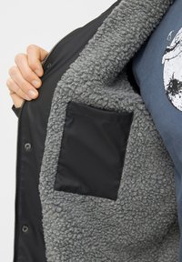 Giacca grigia foderata in shearling con un esterno nero, dotata di chiusura con zip e una tasca nera opaca all'interno della fodera.