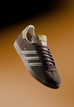 Brązowy but sportowy Adidas Gazelle z białymi paskami, sznurowadłami i podeszwą, przedstawiony na tle o gradiencie brązu.