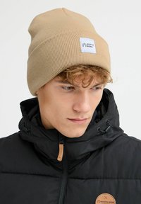 Beige ribbestrikket lue med et hvitt merke med "North Bend"-logo. Kombinert med en svart glidelåsjakke med skinnaksent.