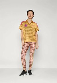 Γυναίκα που στέκεται φορώντας ένα ταιριαστό μοτίβο κίτρινο και ροζ μπλουζάκι κοντό μανίκι και κολάν, μαύρα αθλητικά παπούτσια, σε απλό ανοιχτό γκρίζο φόντο.