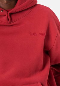 Sudadera roja de tejido suave con un logo bordado de "Sixth June" en el frente. El diseño incluye una capucha con cordón y un bolsillo canguro.