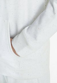 La mano de una persona descansa dentro del bolsillo frontal de un sudadera blanca moteada con pantalones a juego, destacando la suave textura de la tela.