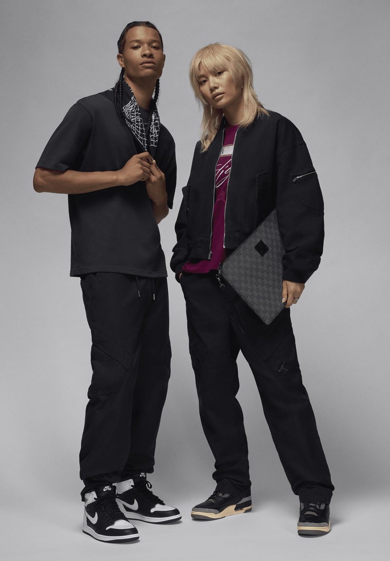 Deux individus se tiennent côte à côte, vêtus de tenues noires assorties. L'homme porte une chemise à manches courtes et un pantalon cargo. La femme arbore une veste bomber noire sur une chemise bordeaux et tient un sac noir texturé. Tous deux portent des baskets au design distinct.
