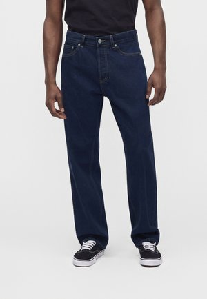 Hombre vestido con vaqueros azul oscuro de pernera recta y zapatillas negras con cordones blancos, de pie frente a un fondo claro y liso.