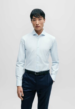 Chemise bleu clair à boutons avec un motif texturé, manches longues et col pointu, associée à un pantalon bleu marine et une ceinture noire.
