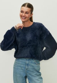 Maglione peloso blu navy con scollatura rotonda e maniche lunghe e voluminose; abbinato a jeans di denim azzurro. Tessuto morbido e dal texture.