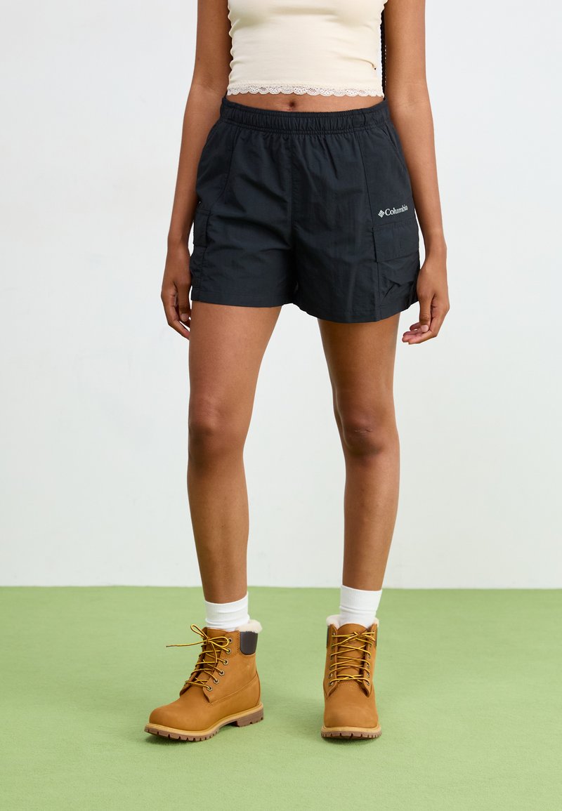 Pantaloni corti neri con vita elasticizzata e tasche laterali, abbinati a stivali color sabbia con lacci. Calze bianche corte visibili sopra gli stivali.