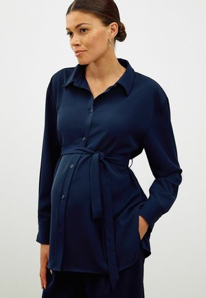 Marinblå button-down skjorta med knyt i midjan, långa ärmar och en slät textur. Har en klassisk krage och sidfickor.
