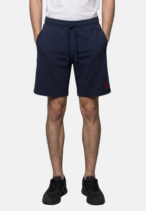 Mann trägt marineblaue Shorts mit Kordelzug und Seitentaschen sowie schwarze Sneaker und steht vor einem einfarbigen Hintergrund.
