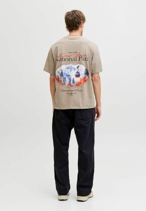 Mann trägt beige T-Shirt mit Seilbahn- und Bergdesign des Great Basin Nationalparks auf dem Rücken, schwarze Hose und helle Sneakers.
