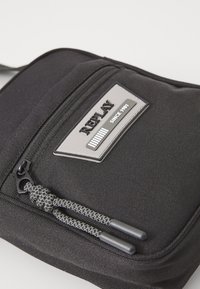 Black fabric crossbody bag featuring a textured front with a zipper, logo patch, and braided zipper pulls. Compact and durable design.