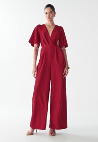 Vrouw in een dieprode wijde jumpsuit met fladdermouwen, een diepe V-hals, een gouden armband en naakthoge hakken op een witte achtergrond.