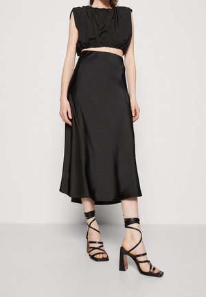 Femme portant un crop-top noir sans manches, une jupe midi assortie et des sandales noires à talons hauts à lanières, debout sur un sol gris.