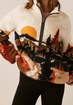 Frau trägt einen Fleece-Pullover mit halbem Reißverschluss, der ein Landschaftsmotiv mit Bäumen, Bergen und einer orangefarbenen Sonne zeigt.
