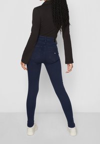 Jeans ajustados de tiro alto en azul oscuro con dos bolsillos traseros, combinados con una blusa negra de manga larga ajustada y zapatillas blancas.