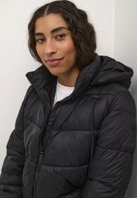 Veste coupe-vent noire avec capuche, dotée d'une fermeture éclair, de cordons de serrage et d'une finition texturée. Portée sur une chemise blanche.