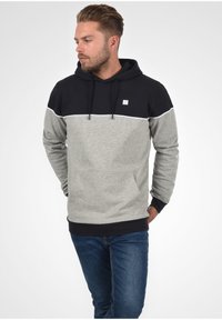 Hoodie met een zwart bovenste gedeelte en een grijs ondergedeelte, voorzien van een voorzak, een witte accentlijn en een klein logo op de borst.