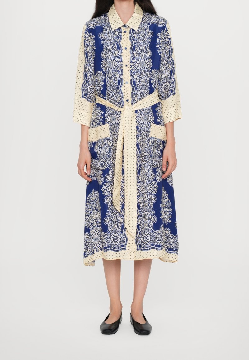 Femme portant une robe à manches longues bleu et crème avec des motifs floraux et à pois, une ceinture à la taille et des chaussures plates noires.