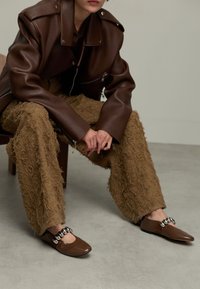 Personne assise portant une veste en cuir marron foncé, un pantalon marron texturé et des chaussures plates marron avec des embellissements ronds métalliques.