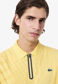 Polo giallo con colletto con zip, caratterizzato da un motivo testurizzato e un logo in evidenza sul petto. Finitura di tessuto liscia.