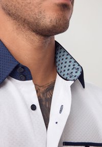 Camicia bianca con superficie strutturata, colletto blu con motivo e bottoni blu scuro. Il colletto presenta disegni geometrici e bordatura a contrasto.