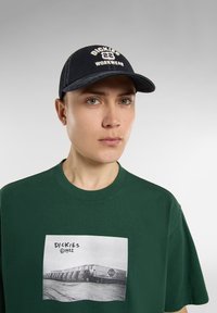 Svart keps med vit broderad "DICKIES 22 WORKWEAR", ihop med en grön t-shirt med en svart och vit grafik av lastbilar.