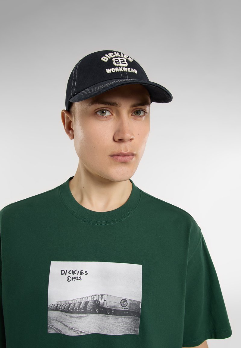 Svart keps med vit broderad "DICKIES 22 WORKWEAR", ihop med en grön t-shirt med en svart och vit grafik av lastbilar.