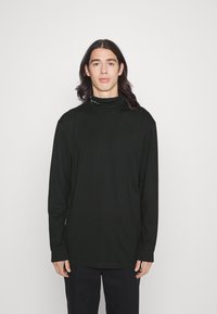 Langärmliges schwarzes Rollkragenshirt aus weichem Stoff mit hohem Kragen und subtiler Markenkennzeichnung am Halsausschnitt, kombiniert mit dunklen Hosen.