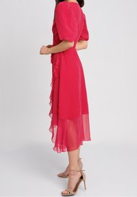 Frau in rotem Midikleid mit weißen Punkten und Puffärmeln, trägt beigefarbene High-Heel-Sandaletten mit Knöchelriemen und posiert seitlich.