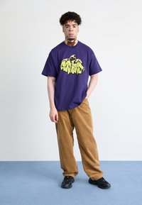 Purple cotton t-shirt with yellow graffiti-style logo, paired with brown corduroy pants, and black sneakers. Model stands on blue carpet.