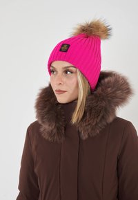 Donna che indossa un berretto lavorato a maglia di un vivace colore rosa con pom-pom in pelliccia e un cappotto marrone con un ampio colletto di pelliccia, che guarda a sinistra su uno sfondo bianco.