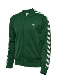 Vihreä vetoketjullinen urheilutakki, jossa valkoiset kalanruotokuvioiset raidat hihoissa ja pieni valkoinen mehiläislogo vasemmassa rinnassa, resorit hihansuissa ja helmassa.