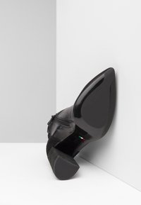 Chaussure à talons hauts en cuir noir avec une finition lisse, un bout pointu, des accents texturés et un détail de fermeture éclair sur le côté. Les semelles sont élégantes et brillantes.