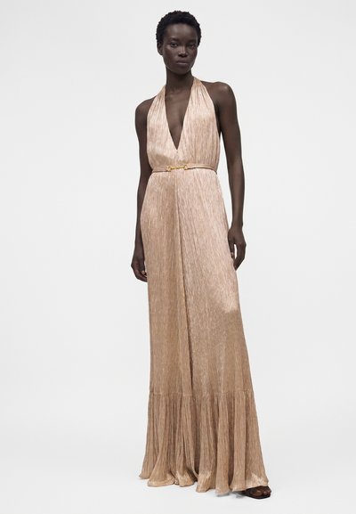 Γυναίκα που φοράει αμάνικο λαμπερό χρυσό φόρεμα με halter λαιμόκοψη, βαθύ V-ντεκολτέ και φαρδύ τελείωμα, στέκεται μπροστά σε μονόχρωμο φόντο.