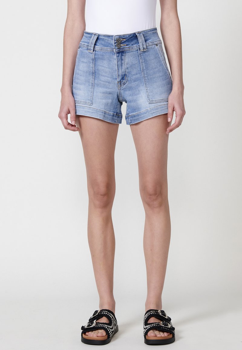 Šviesiai mėlyni džinsiniai šortai, pasižymintys aukštu juosmeniu, dviem priekinėmis kišenėmis ir pažemintu kraštu. Sujungti su juodais sandalais su dekoratyviniais akcentais.