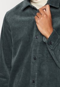 Persona che indossa una camicia a maniche lunghe in velluto a coste verde scuro sopra un dolcevita beige, mentre aggiusta il colletto.