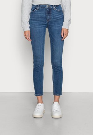 Persoon die blauwe slim-fit jeans draagt, een lichtgrijze trui en witte sneakers, staande op een grijze vloer tegen een lichte achtergrond.