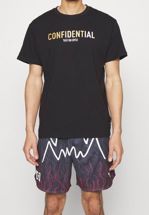 Mies, joka pitää mustaa T-paitaa, jossa on tekstit "CONFIDENTIAL" ja ylösalaisin oleva "FEAR NO EVIL", yhdistettynä mustiin ja punaisiin liehukuvioisiin shortseihin.