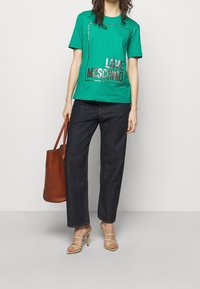 Kvinna i en grön "Love Moschino" t-shirt, mörkblå jeans, beige klacksandaler, håller en stor brun läderhandväska.