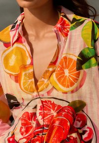 Camicia button-up colorata con un vivace motivo di frutta, arricchita da arance, limoni e accenti rossi su uno sfondo a righe rosa chiaro.