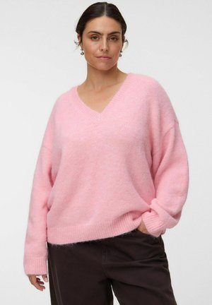 Femme portant un pull rose ample à col en V et un pantalon foncé, debout avec une main dans la poche, devant un fond clair uni.