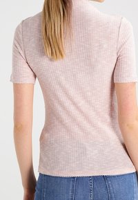 Světle růžový, krátký rukáv, top vyrobený z ribbed látky s vysokým výstřihem a přiléhavým střihem. Okraje mají surový, nedokončený vzhled.