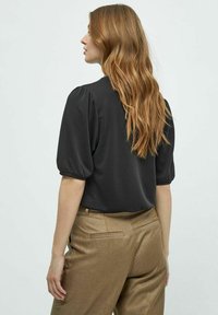 Zwarte blouse met pofmouwen, gemaakt van een gladde stof, gecombineerd met hoge, gestructureerde gouden-brown broek, gezien vanuit de achterkant.