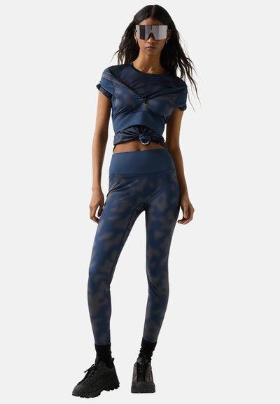 Frau trägt reflektierende, übergroße Sonnenbrille, marineblaue Leggings mit Tarnmuster, passendes kurz geschnittenes Oberteil, das an der Taille gebunden ist, und schwarze Sneakers.