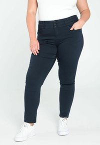 Jean skinny bleu foncé avec fermeture à trois boutons et poches latérales, en denim extensible, associé à des baskets blanches. Texture lisse.
